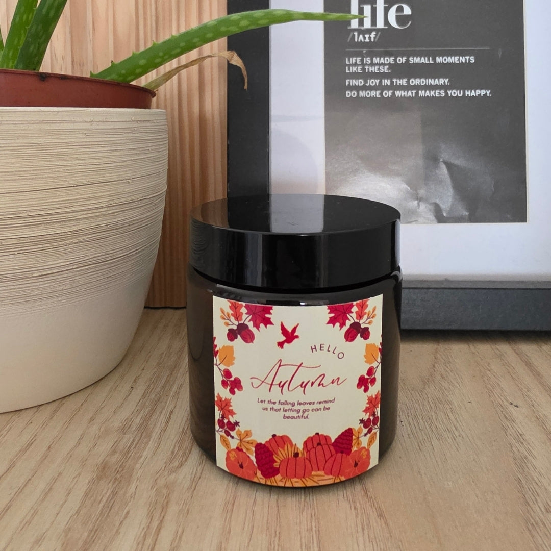 Świeca sojowa Pumpkin Spice w brązowym słoiczku, zamknięta czarną nakrętką, z etykietą „Hello Autumn” z jesiennym wieńcem kwiatowym, na drewnianej półce, obok aloes w doniczce i oprawiony obrazek
