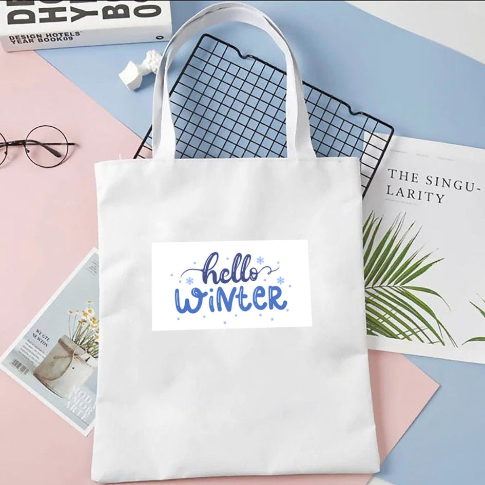 Torba płócienna Kiskedves Hello Winter w kolorze białym, z niebieskim napisem hello winter, na pastelowym tle obok magazynów i okularów.