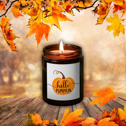 Płonąca świeca sojowa Pumpkin Spice w brązowym słoiczku z etykietą „hello PUMPKIN”, wśród jesiennych liści na drewnianym stole