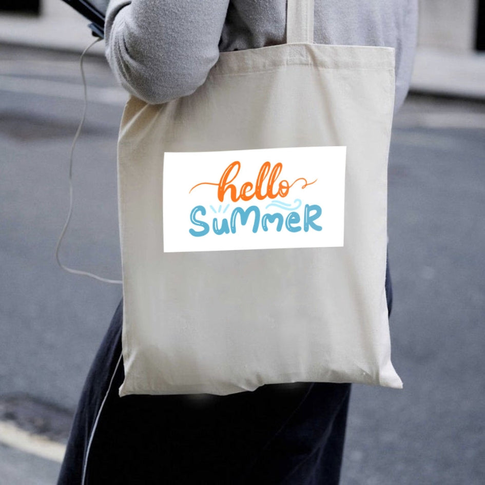 Naturalna torba płócienna Kiskedves Hello Summer noszona na ramieniu, z pomarańczowo-niebieskim napisem hello summer, w miejskim otoczeniu.