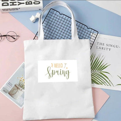 Torba płócienna Kiskedves Hello Spring w kolorze białym, z zielono-pomarańczowym napisem hello spring, na pastelowym tle obok magazynów.