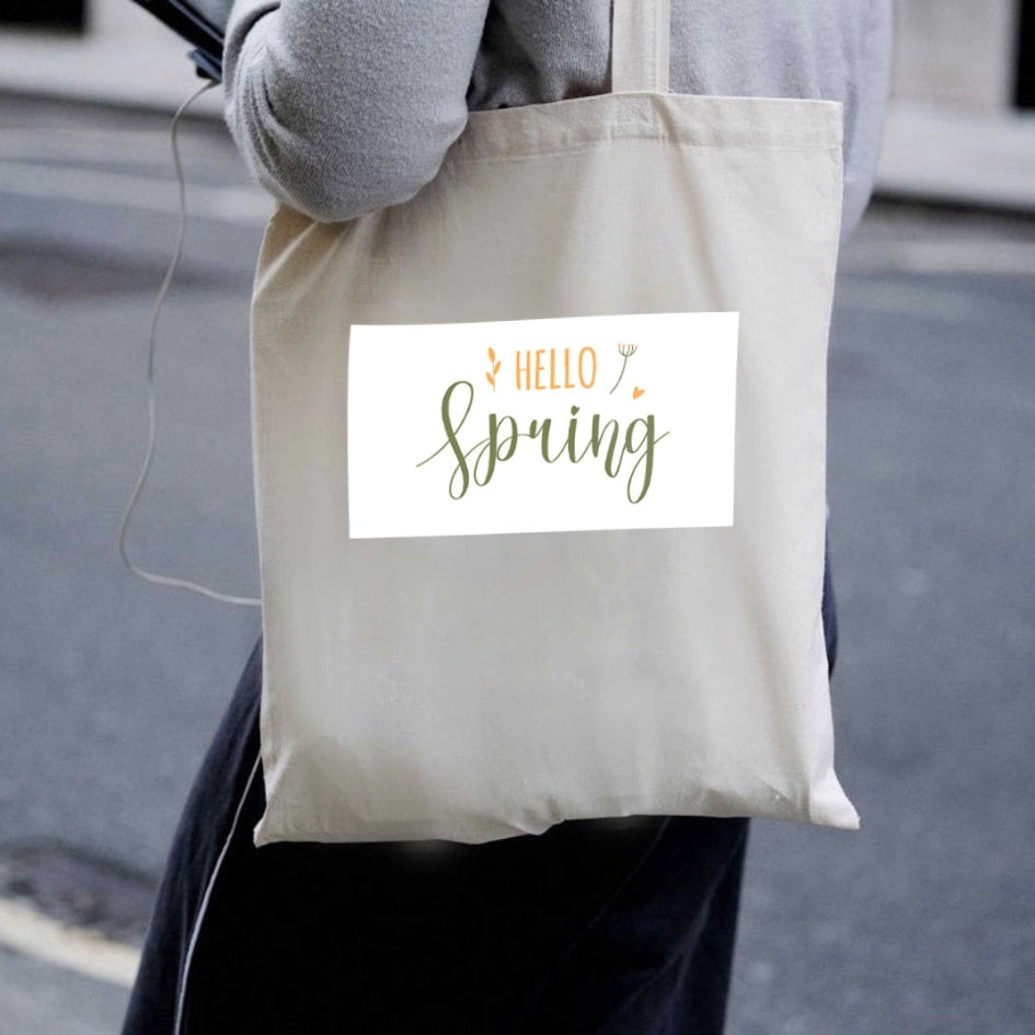 Naturalna torba płócienna Kiskedves Hello Spring noszona na ramieniu, z zielono-pomarańczowym napisem hello spring, w miejskim otoczeniu.