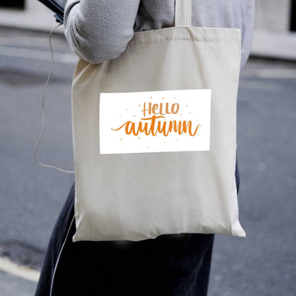 Naturalna torba płócienna Kiskedves Hello Autumn noszona na ramieniu, z pomarańczowym napisem hello autumn, w miejskim otoczeniu.