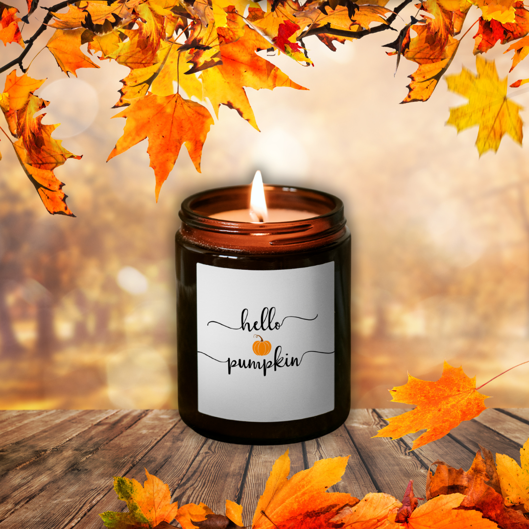 Płonąca świeca sojowa Pumpkin Spice w brązowym słoiczku z etykietą „hello pumpkin”, wśród jesiennych liści na drewnianym stole