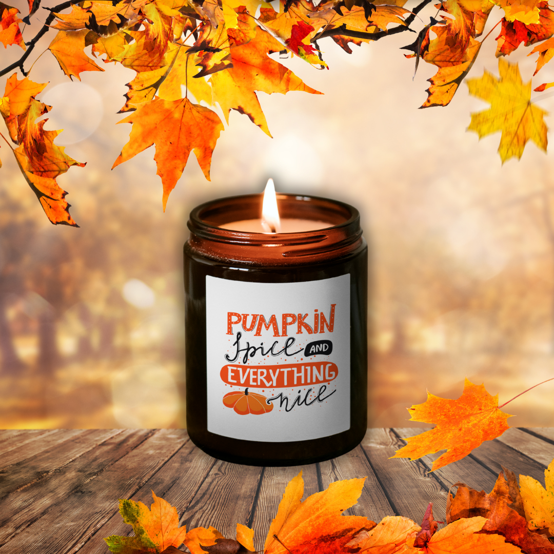 Płonąca świeca sojowa Pumpkin Spice w brązowym słoiczku z etykietą „Pumpkin Spice and everything nice”, wśród jesiennych liści na drewnianym stole