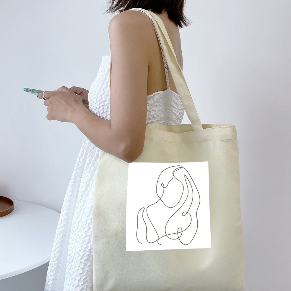 Torba płócienna Kiskedves Line Art na ramieniu, ze splecionymi postaciami, do białej letniej sukienki.