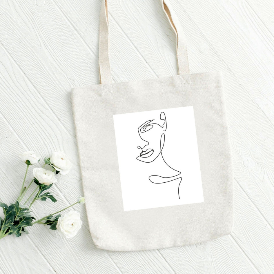 Torba płócienna Kiskedves Line Art z kobiecym profilem, na białych deskach obok białych jaskrów.