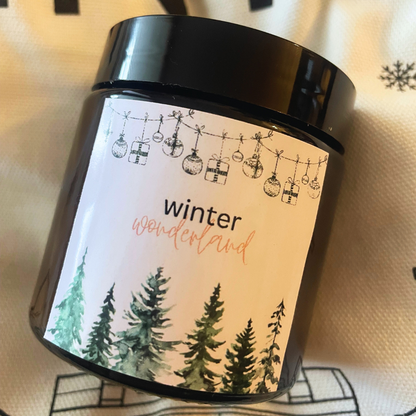 Świeca Kiskedves Winter Wonderland na tkaninie w płatki śniegu, z bliska widać etykietę z lasem.