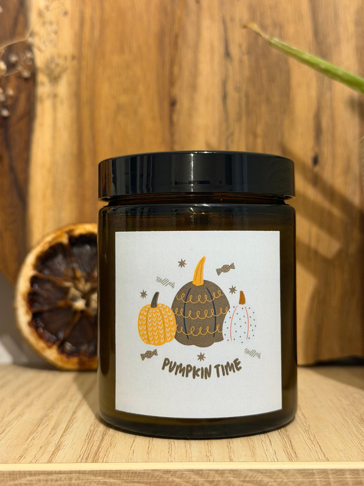 Świeca sojowa Pumpkin Spice w brązowym słoiczku, zamknięta czarną nakrętką, z etykietą „Pumpkin time” z rysowanymi dyniami, na drewnianej półce, obok suszony plaster pomarańczy