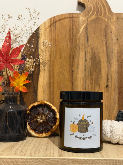 Świeca sojowa Pumpkin Spice w brązowym słoiczku z etykietą „Pumpkin time”, przed drewnianą deską w kształcie dyni, obok wazon z suszonymi kwiatami, jesienne liście i biała szydełkowa dynia