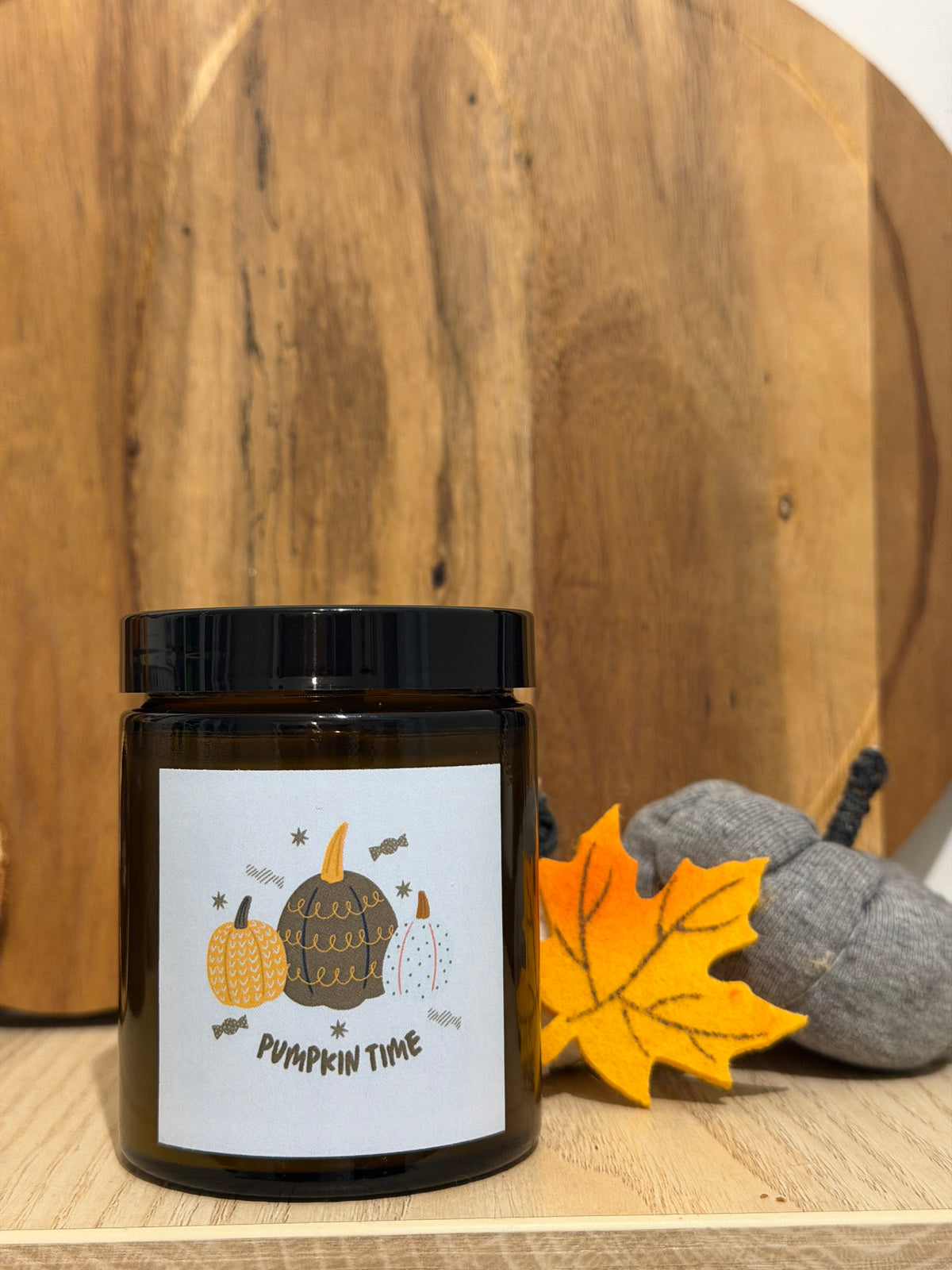 Świeca sojowa Pumpkin Spice w brązowym słoiczku z etykietą „Pumpkin time”, przed okrągłą drewnianą deską, obok żółty filcowy liść klonu i szare materiałowe dynie