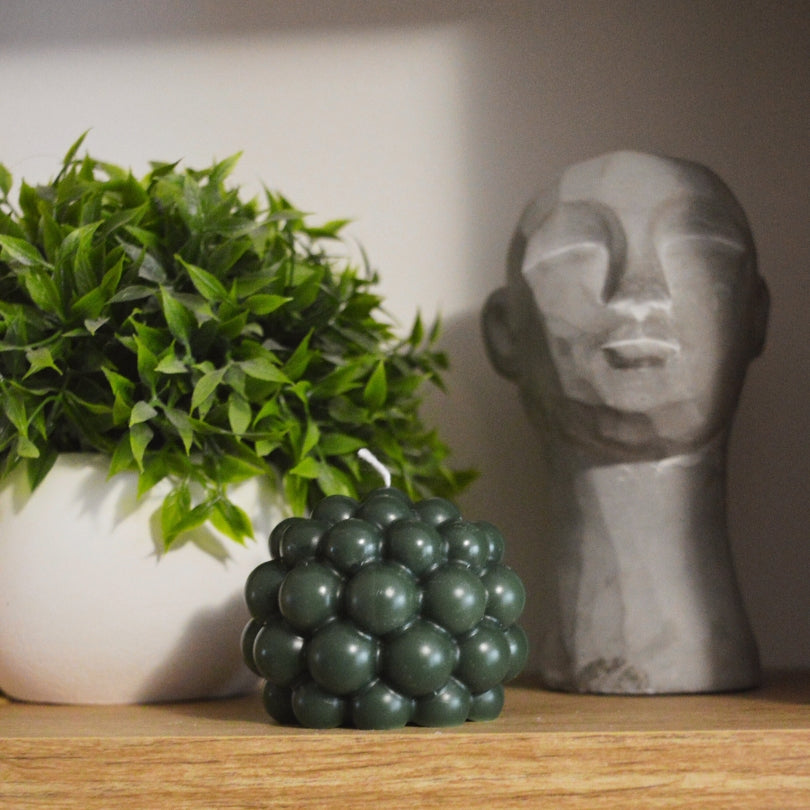 Świeca Kiskedves Bubi w zieleni: ciemnozielona bąbelkowa kostka na drewnianej półce obok rośliny.
