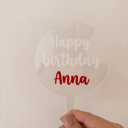 Przezroczysty topper z akrylu z białym napisem „Happy birthday” i czerwonym imieniem „Anna”, trzymany w dłoni