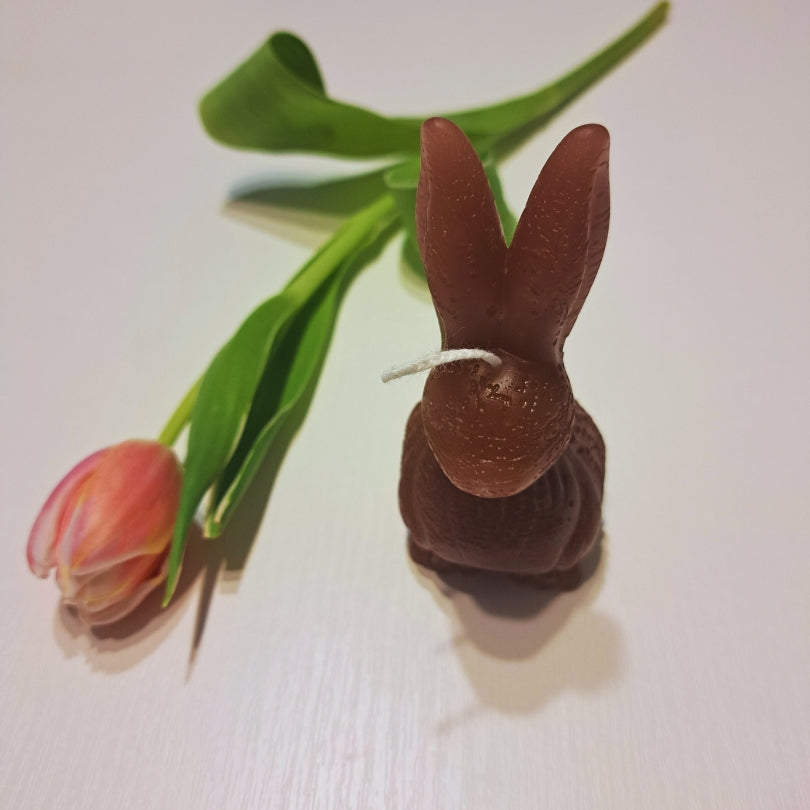 Brązowa świeca Kiskedves w kształcie króliczka z długimi uszami na białym drewnianym stole, obok różowy tulipan.