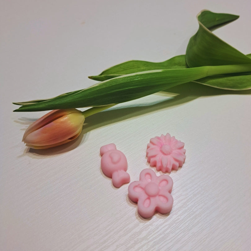 Trzy różowe woski zapachowe Kiskedves – króliczek, margerytka i kwiat – obok tulipana, na białej drewnianej powierzchni.