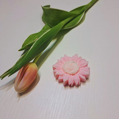 Różowy wosk zapachowy Kiskedves w kształcie kwiatu na białym drewnianym stole, obok różowawy tulipan i zielone liście.