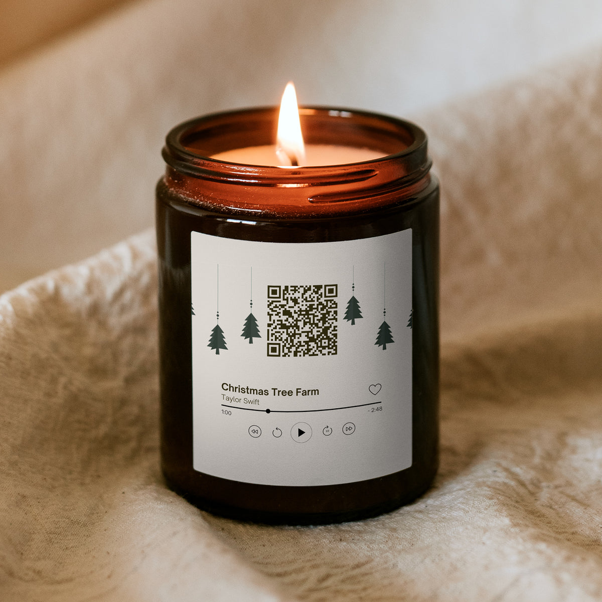 Płonąca świąteczna świeca „Melodyjna” Kiskedves w brązowym słoiczku na miękkim kocu, na etykiecie kod QR i karta odtwarzacza z piosenką Christmas Tree Farm