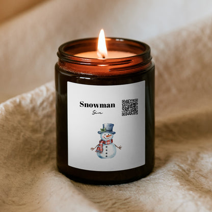 Płonąca świąteczna świeca „Melodyjna” Kiskedves w brązowym słoiczku na miękkim kocu, na etykiecie kod QR, napis Snowman i akwarelowy bałwan